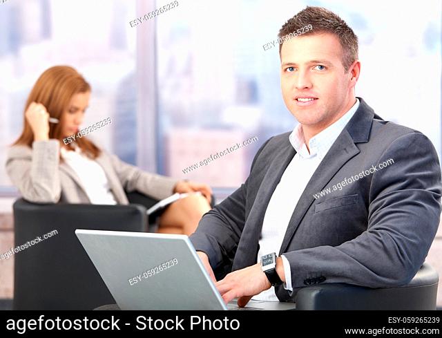 Middle-aged businessman using laptop, sitting in office lobby, having conference break, smiling