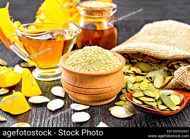 Pumpkin flour in bowl, oil in sauceboat and jar, seeds in bag, on table and in spoon, slices of vegetable on wooden board background
