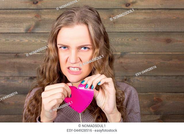 Young girl with angry face holding pink heart on a stick in her hands, wooden background. Fun photo props and accessories for shoots