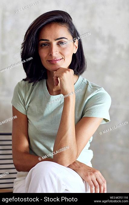 Close-up of smiling businesswoman with hand on chin sitting against wall in office