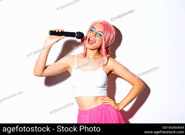 Portrait of joyful beautiful female in pink wig, with bright makeup, singing karaoke in microphone, standing over white background
