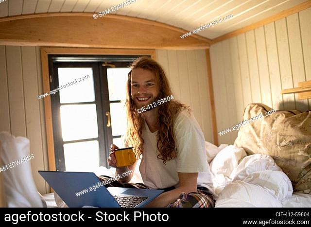 Portrait happy young man with coffee and laptop in bed