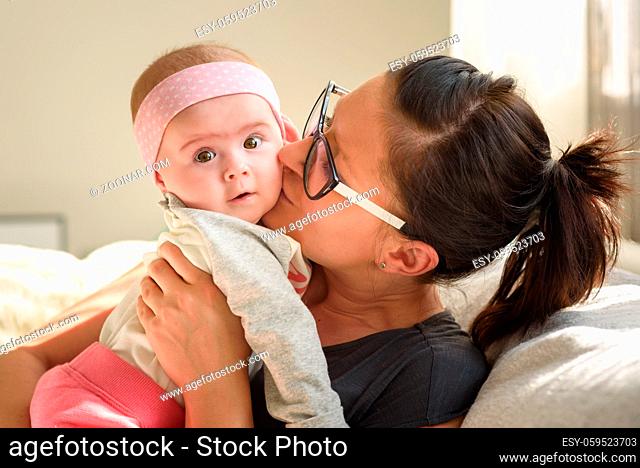 Mother holding and kissing beautiful baby girl in bright sunny room. Motherhood background