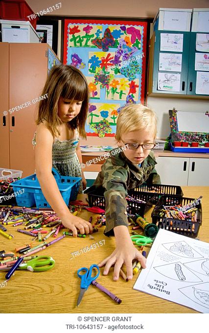 Kindergarten children in San Clemente, CA, collect and organize a table full of classroom objects including crayons and worksheets