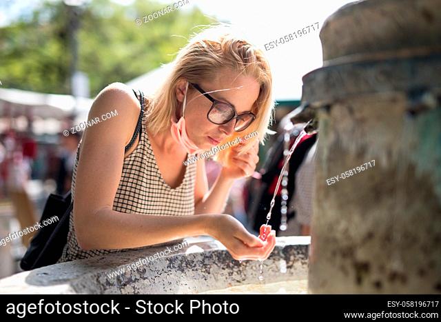 Thirsty young casual cucasian woman wearing medical face mask drinking water from public city fountain on a hot summer day