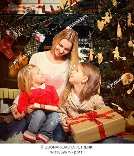 Young mother and her two little daughters with Christmas gifts by a Christmas tree in cozy living room in winter