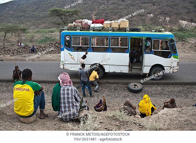 A bus employee is changing the flat tyre of the bus he is working in. The passengers are watching the young man changing the tyre