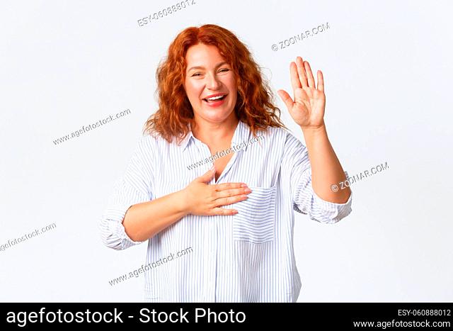 Portrait of cheerful honest, smiling middle-aged redhead woman, mother making promise, raising one arm and hold hand on heart as pleading, give her word or oath