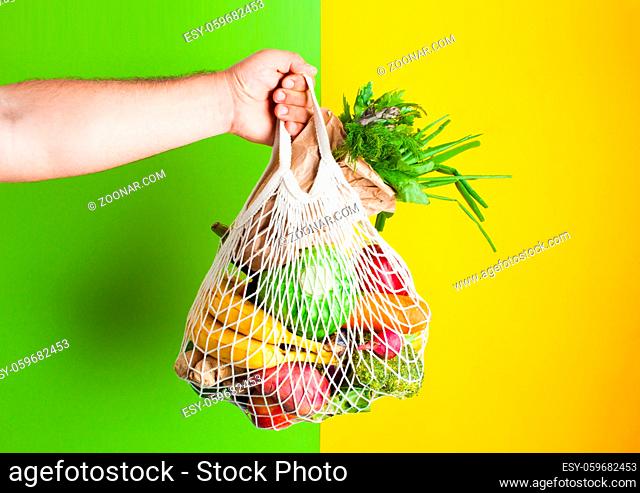 Zero waste shopping. Man hand holds cotton bag full of vegetables on green and yellow background. Produce in paper bag. Place for text, vegan food