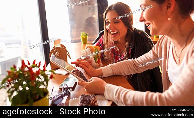 Toned picture of best friends ladies spending free time in cafe. One girl using tablet PC while another eating delicious sandwich or snack