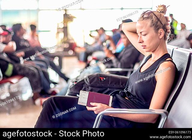 Casual tanned blond female traveler holding cell phone, passport and boarding pass while waiting to board a plane at the departure gates at the asian airport...