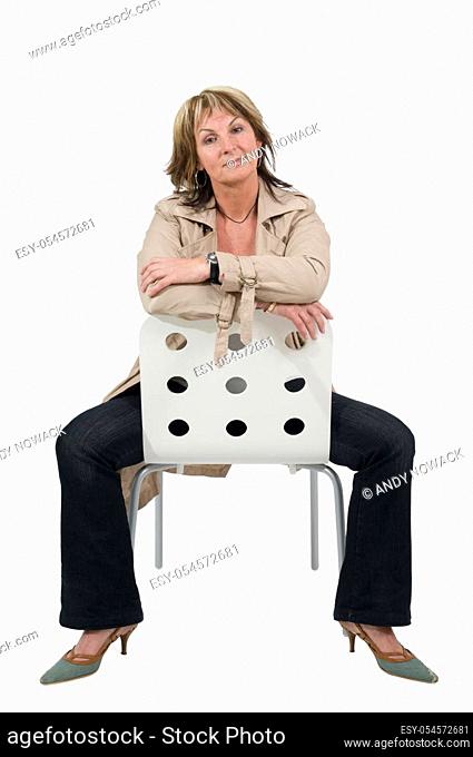 Full body portrait of a mature woman in a trench coat and black pants sitting astride a white chair with her forearms on top of each other against the white...
