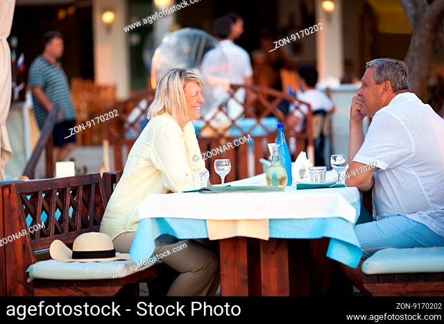 Middle-aged couple seated at a table in a restaurant enjoying a meal and wine while they smile and chat, side view with people visible in the background