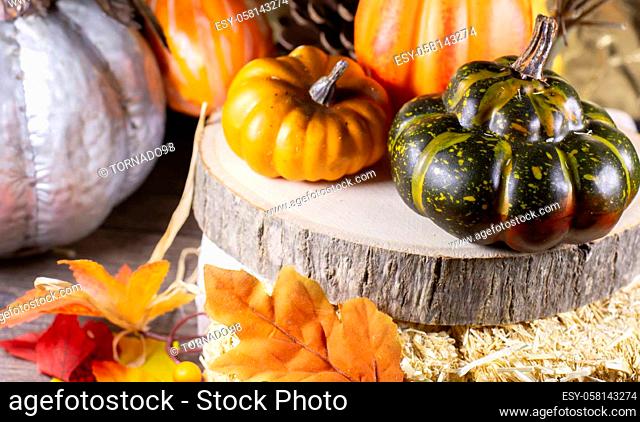 Small orange pumpkins, orange squash, pinecones, and green squash on wood stacked on leaf-covered hay with a golden background, next to a silver pumpkin nearby