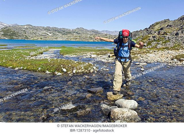 Young woman crossing a creek, hiking, backpacking, hiker with backpack, historic Chilkoot Trail, Chilkoot Pass, Crater Lake behind, alpine tundra