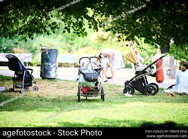 25 June 2022, Berlin: Baby carriages stand in the shade of the trees while parents play with their children at the watering hole in Volkspark am Weinberg
