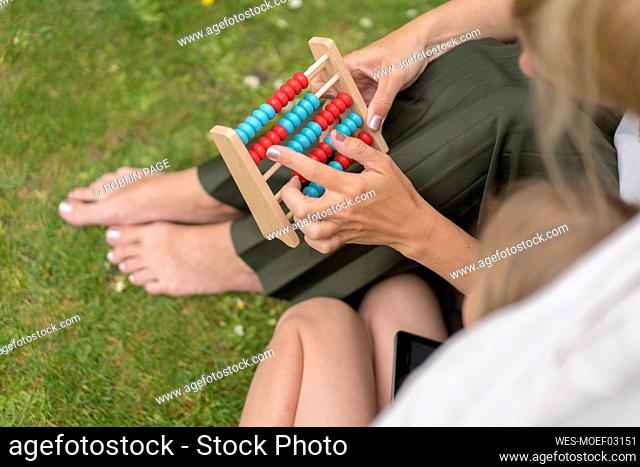 Mother teaching abacus to daughter while sitting on grass at back yard during weekend