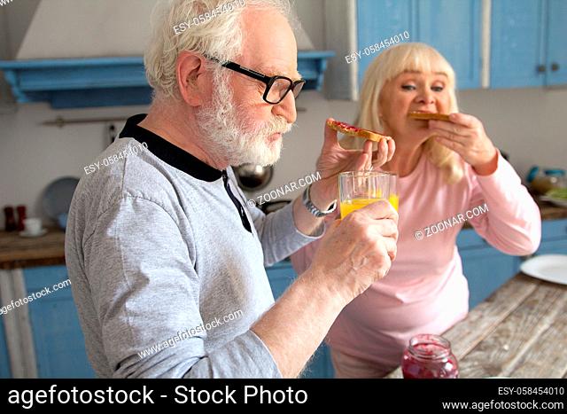 Old grandpa chewing on some jam toasts with his wife at breakfast. Cute old man with glasses and white beard having some bread with jelly and orange juice for...