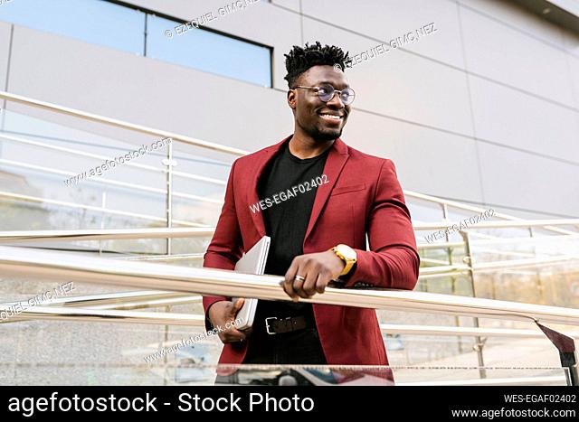 Smiling male professional holding laptop while standing by railing