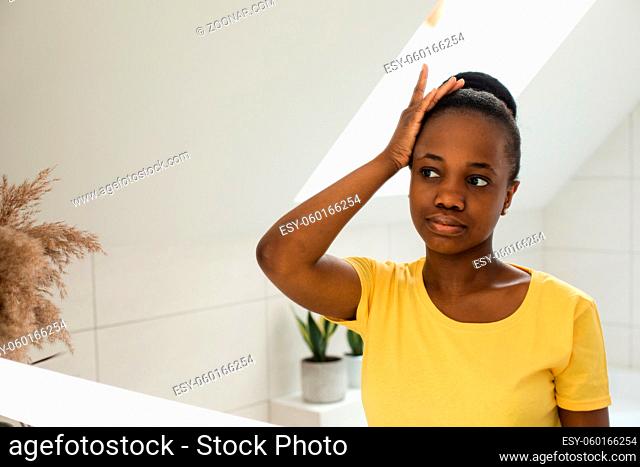 Young charming Afro-American girl brushing her hair while looking into a mirror. Everyday hair care routines. Young woman with brush making order of messy hair