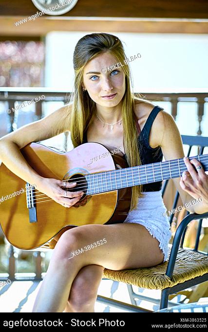 Portrait of a young beautiful caucasian woman in her 20's with long hair and blue eyes playing guitar on the porch of a country house