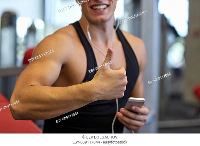 sport, bodybuilding, lifestyle, technology and people concept - happy man with smartphone and earphones listening to music and showing thumbs up in gym