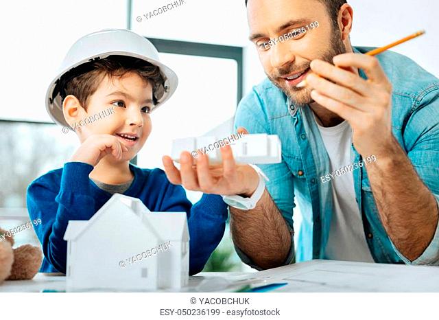 Checking results. Upbeat father and his little son sitting at the table in the office and looking at the results of 3D printing, being the part of a house model