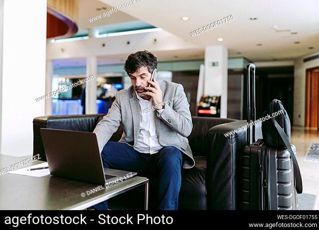 Businessman talking on phone while using laptop in hotel lobby