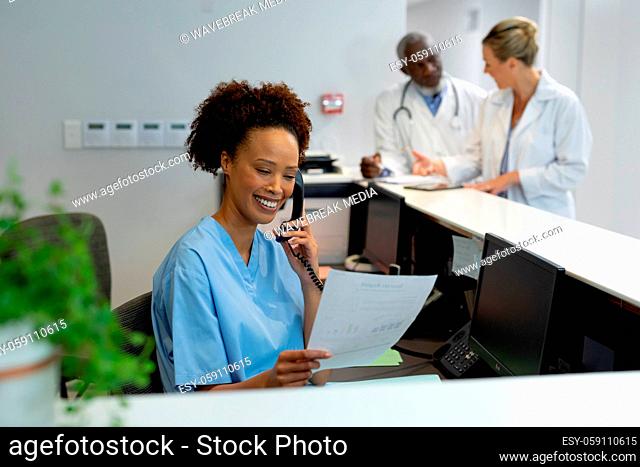 Mixed race female doctor at desk in hospital reception talking on phone and holding document