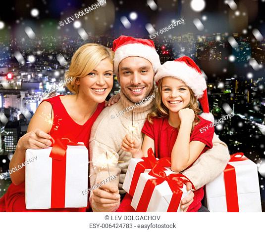 christmas, holidays, family and people concept - happy mother, father and little girl in santa helper hats with gift boxes over snowy night city background