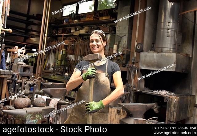 27 July 2023, Saxony, Hohenprießnitz: The 39-year-old blacksmith and metal designer Marika Widdermann stands laughing with a sledgehammer in the forge of her...