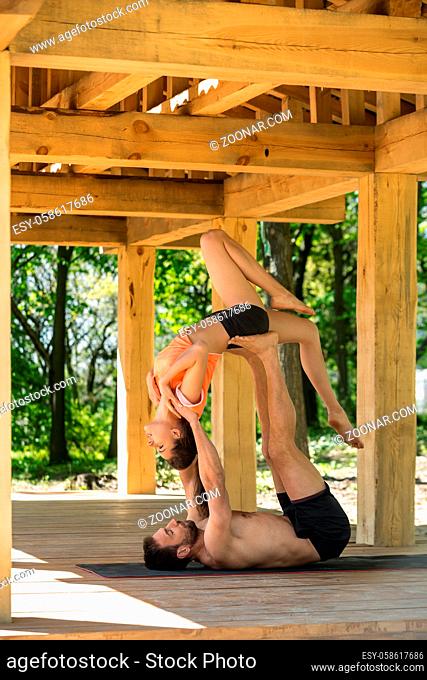 Pretty couple are engaged in yoga on the wooden terrace on the nature background. Man lies on his back on the black yoga mat and holds the girl in the air with...