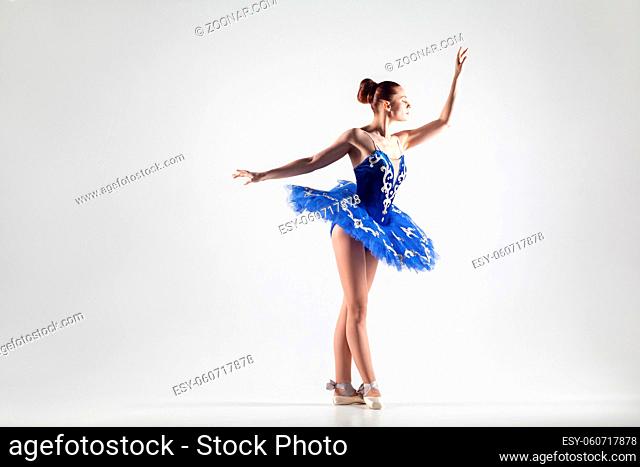 attractive ballerina with bun collected hair wearing blue dress and pointe shoes performing in white studio. indoor, studio shot