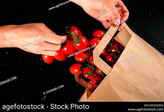 Woman hand taking fresh cherry tomatoes with green vines from brown paper shopping bag over black table, close up detail