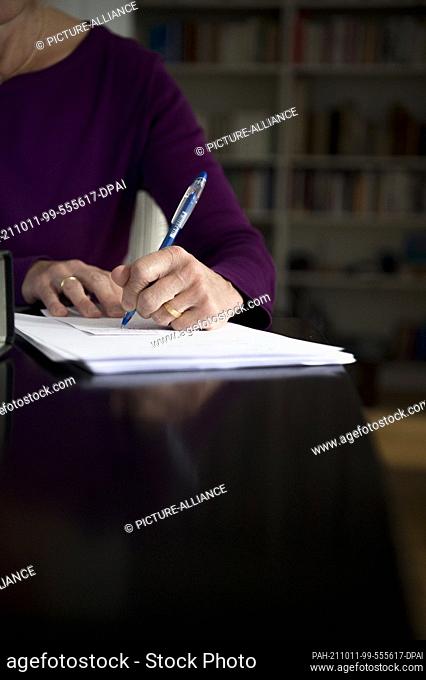 PRODUCTION - 01 October 2021, Bavaria, Munich: A woman is sitting at a dining table at home. She is writing with a pen on a piece of paper