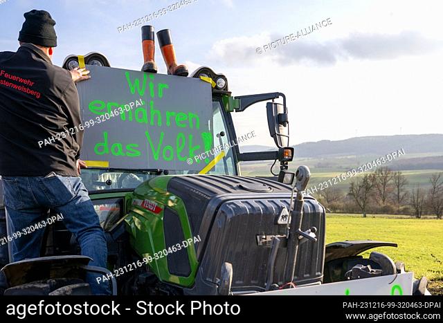 16 December 2023, Bavaria, Wüstenwelsberg: A farmer attaches a protest sign to his tractor. German farmers are up in arms against the announced abolition of the...