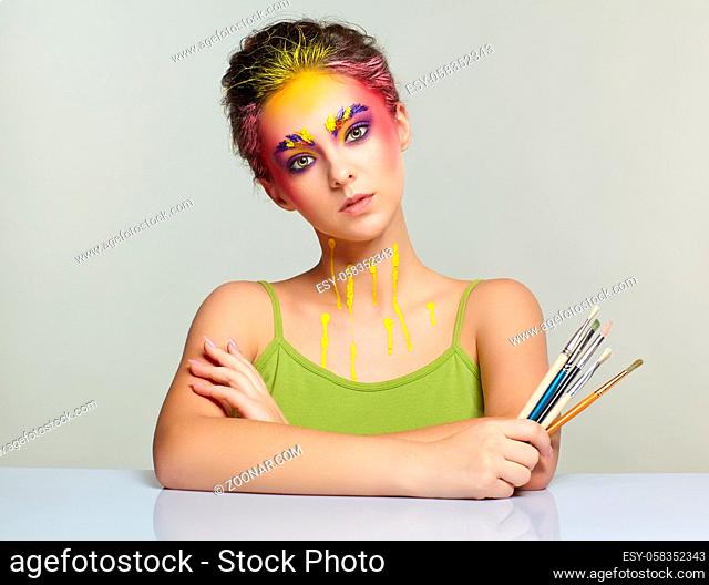 Portrait of young woman posing at the table with brushes in hand. Unusual female art make-up with paint on brows, hair and around eyes
