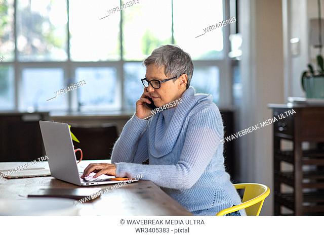 Side view close up of a mature Caucasian woman with short grey hair wearing glasses sitting at her dining room table making a phone call on a smartphone and...