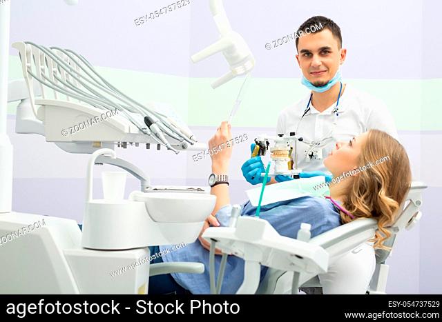 Young dentist working with the patient in a modern hospital. He is assisted by a young girl assistant. The patient sees in the mirror the result of the work of...
