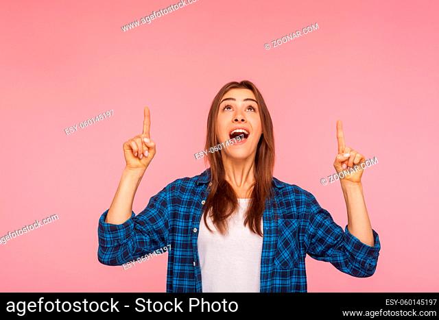 Wow, look above, crazy advertisement. Portrait of amazed brunette girl in checkered shirt pointing up at empty place for ad content and expressing surprise
