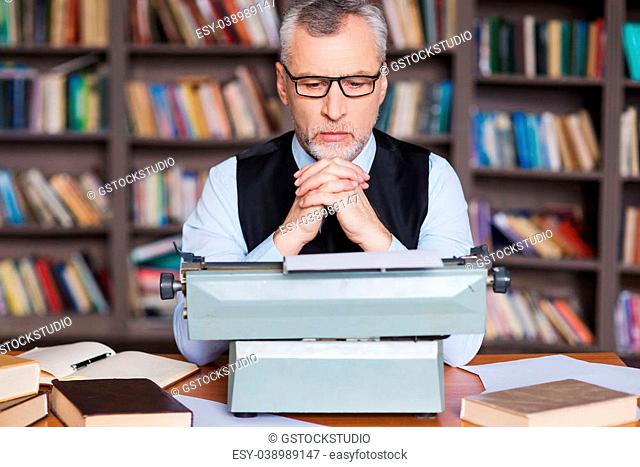 Waiting for inspiration. Confident grey hair senior man in formalwear sitting at the typewriter and looking concentrated with bookshelf in the background