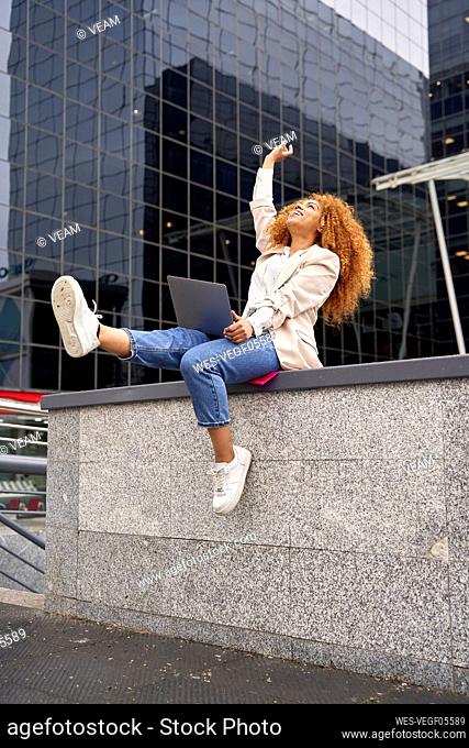 Cheerful businesswoman with laptop looking up sitting on wall outside office building
