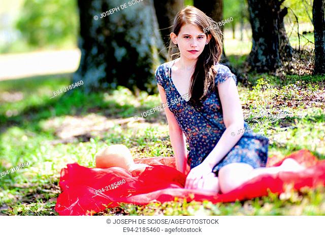 A 22 year old brunette woman wearing a summer dress sitting on a blanket in a pasture