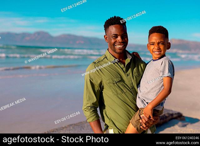 Portrait of african american smiling young man carrying son while standing against sea and blue sky