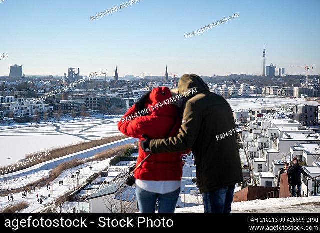 13 February 2021, North Rhine-Westphalia, Dortmund: Day trippers stand on the slag heap above the frozen Phoenix Lake and look out over the panorama of the city...