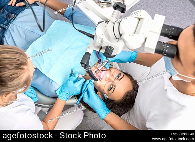 Image of a woman in protective glasses on a patient chair in the dentist's office. Dentist treats her teeth using a dental microscope and instruments