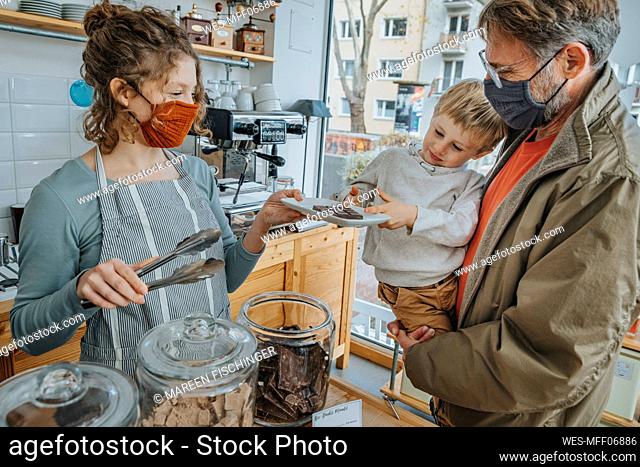 Female owner serving chocolate in plate to little by in candy store during pandemic