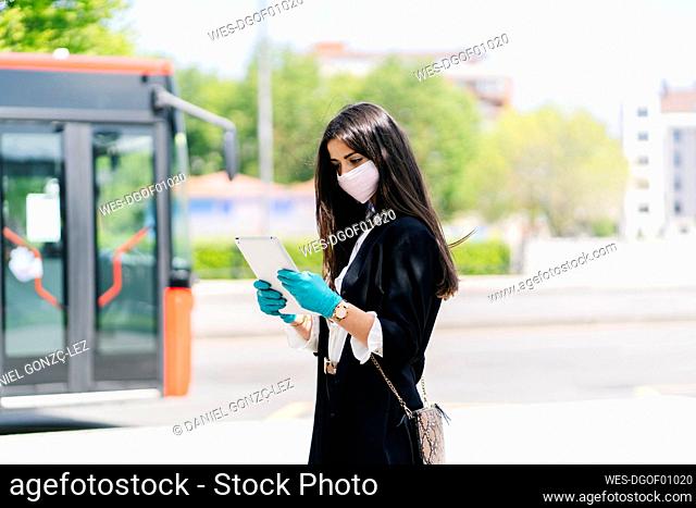 Young woman wearing protective mask and gloves using digital tablet while waiting at bus stop, Spain