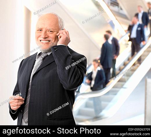 Happy senior businessman standing in office lobby, taking phone call, smiling at camera