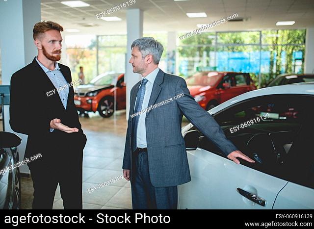 Handsome man at auto showroom talking with vehicle dealer. Car sale, sales manager showing cars to a buyer
